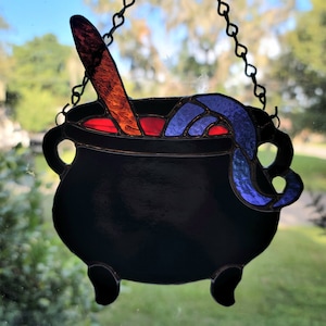May include: A stained glass hanging of a black cauldron with a red and orange stirring spoon and blue and purple smoke rising from it.