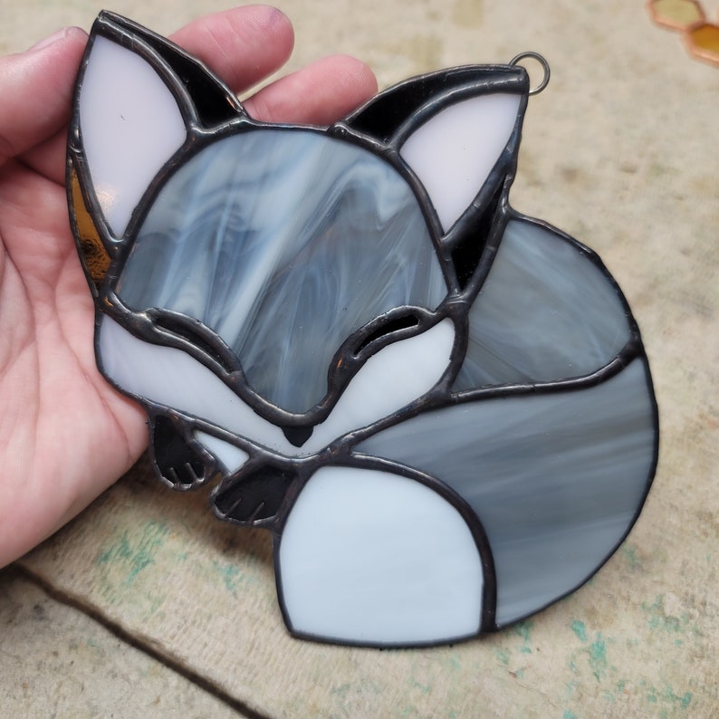 Op de afbeelding: Een gebrandschilderd glazen vosornament in tinten grijs, wit en zwart. De vos is opgerold, met een lus aan de bovenkant om op te hangen. Het glas heeft een gemarmerd effect en de randen zijn zwart omlijnd.