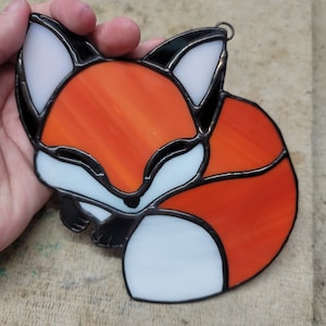 Op de afbeelding: Een gebrandschilderd glazen vosornament met oranje, witte en zwarte secties. De vos is opgerold, met een zwarte omtrek en een metalen lus om op te hangen. Het glas heeft een getextureerd uiterlijk.