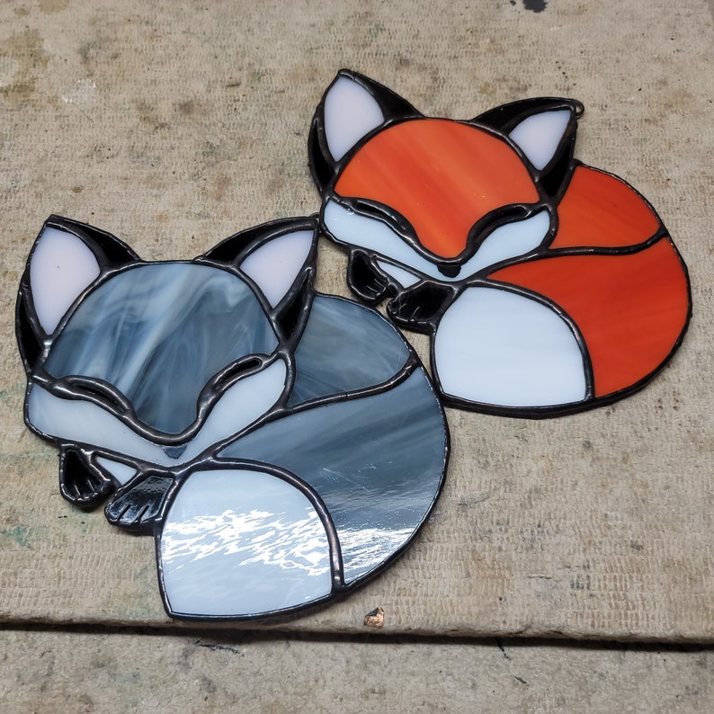 Op de afbeelding: Twee glas-in-lood vossen ornamenten. E&eacute;n vos is oranje, wit en zwart. De andere is grijs, wit en zwart. Beide zijn opgerold, met gedetailleerd glaswerk en een metalen rand.