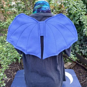 Puede incluir: Accesorio de disfraz de alas de fieltro azul. Las alas están unidas a una camiseta negra. Las alas tienen una forma curva con detalles cosidos. La persona lleva un sombrero colorido. El fondo es un jardín.