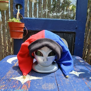 May include: A jester hat made of fleece fabric featuring a split design with one side in bright red and the other in royal blue. The hat has a wide black brim and two long points, each finished with a small metallic bell at the tip, displayed on a silver alien-shaped mannequin head.