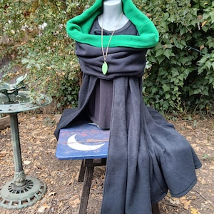 Puede incluir: Una capa de forro polar gris oscuro con un forro interior verde vibrante y una capucha grande. La capa está colocada sobre un maniquí, que también lleva una camiseta gris oscuro sencilla y un collar largo con un colgante en forma de hoja verde.