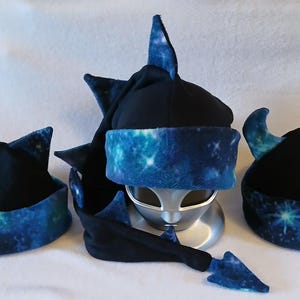 May include: Three novelty hats in a row, each with a different design. The hats are black with a blue and white galaxy print trim. One hat has cat ears, another has a tall fin, and the third has horns.