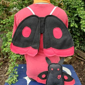 Può includere: Uno zaino con ali di falena nere e rosse con un peluche abbinato. Le ali sono nere con dettagli rossi e un design circolare. Il peluche è nero con dettagli rossi, inclusi occhi e dettagli delle ali. Gli oggetti sono su una superficie blu.