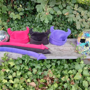 May include: Three fleece beanies with devil horns in hot pink, black, and purple, accompanied by three matching long stuffed tails. A silver disco ball, a small skull figurine, and a faceted iridescent skull are placed on the wooden surface.