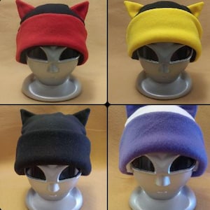 May include: Four fleece beanies with cat ears displayed on silver mannequin heads. The hats feature color combinations of black and red, black and yellow, solid black, and a purple and white striped design with a folded brim on each hat.