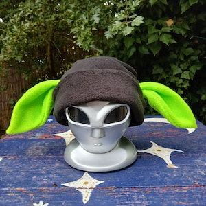 May include: A dark gray fleece beanie featuring two large, pointed, neon green ears extending from the sides. The hat is displayed on a silver-colored, stylized head mannequin with smooth, abstract facial features and dark, oval-shaped eye areas.
