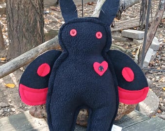 Puede incluir: Una criatura de peluche negra con dos orejas puntiagudas, dos ojos de botón rojos y dos alas grandes con detalles circulares rojos. Un pequeño corazón de tela roja con un botón negro en el centro está sujeto al pecho. El peluche es texturizado.
