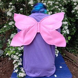 May include: A person wearing a purple shirt and pink butterfly wings. The wings are attached to the back of the shirt. The person is sitting on a blue bench with white floral accents. The background features white flowers and green foliage.