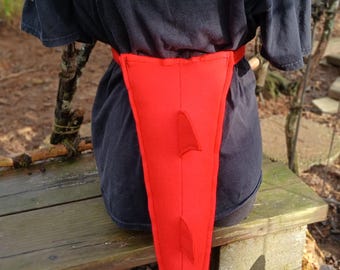 May include: A bright red fabric tail accessory featuring three triangular spikes along the center and a pointed arrow-shaped tip at the end. The tail is attached to a red waistband and is worn over a plain black short-sleeved t-shirt.