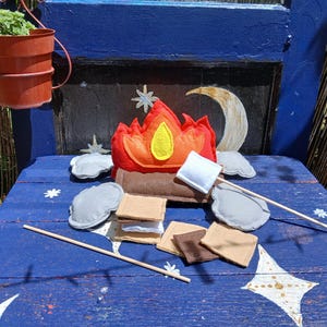 Puede incluir: Un set de fogata de fieltro con una llama roja y naranja, troncos marrones y piedras grises. El conjunto incluye palitos de madera, malvaviscos de fieltro blanco y cuadrados de fieltro marrón y beige que representan componentes de s'mores.