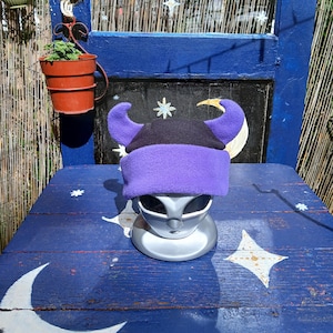 May include: A fleece hat featuring a black crown and a purple brim with two large, curved purple horns extending from the sides. The hat is displayed on a silver-colored mannequin head, showcasing the soft texture and vibrant color contrast.