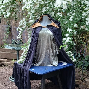 May include: A black velvet cloak with a gold-colored hood and trim, displayed on a mannequin. The cloak is draped over a silver-green dress. The display is set on a blue table with a crescent moon design, against a backdrop of white flowers.