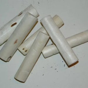May include: A collection of six cylindrical, off-white bone toggles arranged in a scattered pile. Each toggle features a smooth, matte surface with natural variations in colour, including small brown markings and organic textures along the sides.