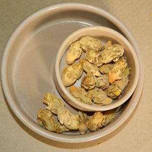 May include: A beige ceramic bowl containing a collection of dried, light-tan flower buds. A smaller, matching beige bowl is nested inside the larger one, also filled with the same dried botanical elements, creating a layered, circular arrangement.