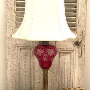 May include: A vintage red glass lamp with an etched design and a white lampshade. The lamp has a brass base and sits on a marble base.