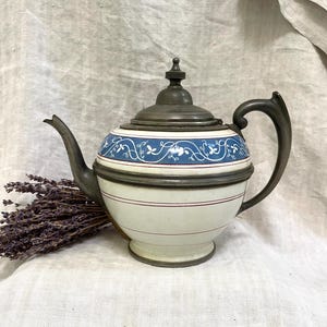 May include: A vintage silver teapot with a white and blue floral design. The teapot has a round body and a spout on the side. The lid is made of silver and has a knob on top.