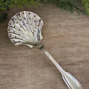 May include: A silver-colored serving spoon with a shell-shaped bowl and a long, slender handle. The bowl has a series of small holes and a ridged design. The handle has a decorative pattern near the bowl and a rounded end.