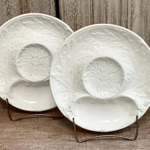 May include: Two white ceramic plates featuring a raised relief pattern of acanthus leaves and a central flower motif. Each plate has a circular indentation in the middle and a smooth, glossy finish, displayed upright on thin gold metal wire stands.