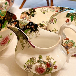 May include: A white porcelain creamer with a floral design in red, green, and yellow. The creamer has a curved spout, a handle, and sits on three small feet. Other floral patterned dishes are in the background.