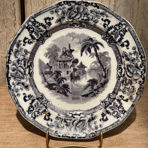 May include: Antique white and blue plate with a detailed landscape scene in the center. The plate features a floral border and a scalloped edge. The plate is displayed on a gold-colored metal stand.