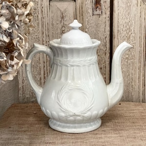 May include: White ceramic teapot with a lid, handle, and spout. The teapot features a decorative design with a wreath and vertical lines. The teapot is sitting on a wooden surface. A dried flower arrangement is in the background.