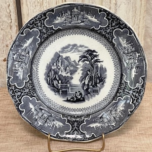 May include: A blue and white decorative plate with a landscape scene in the center. The plate has a scalloped edge and a detailed border with architectural elements. The plate is displayed on a gold-colored metal stand.