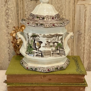 May include: A white ceramic sugar bowl with a lid, featuring a brown and green landscape scene with a bridge and a building. The bowl has a decorative border with red accents and is sitting on top of three green books.