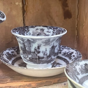 May include: A white teacup and saucer set with a black and white landscape design. The teacup has a flared rim and sits on a matching saucer. The set is likely vintage or antique.