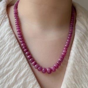 May include: A necklace of polished, round, deep pink gemstones. The beads graduate in size, with the largest at the center and decreasing towards the clasp. The necklace is displayed against a white textured fabric.