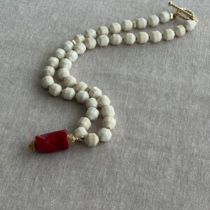 May include: A white beaded necklace with a red coral pendant. The necklace is made of faceted beads and has a gold clasp.