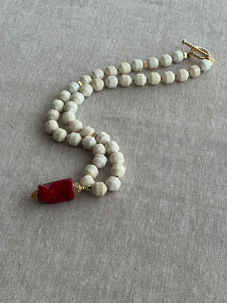 May include: A white beaded necklace with a red coral pendant. The necklace is made of faceted beads and has a gold clasp.