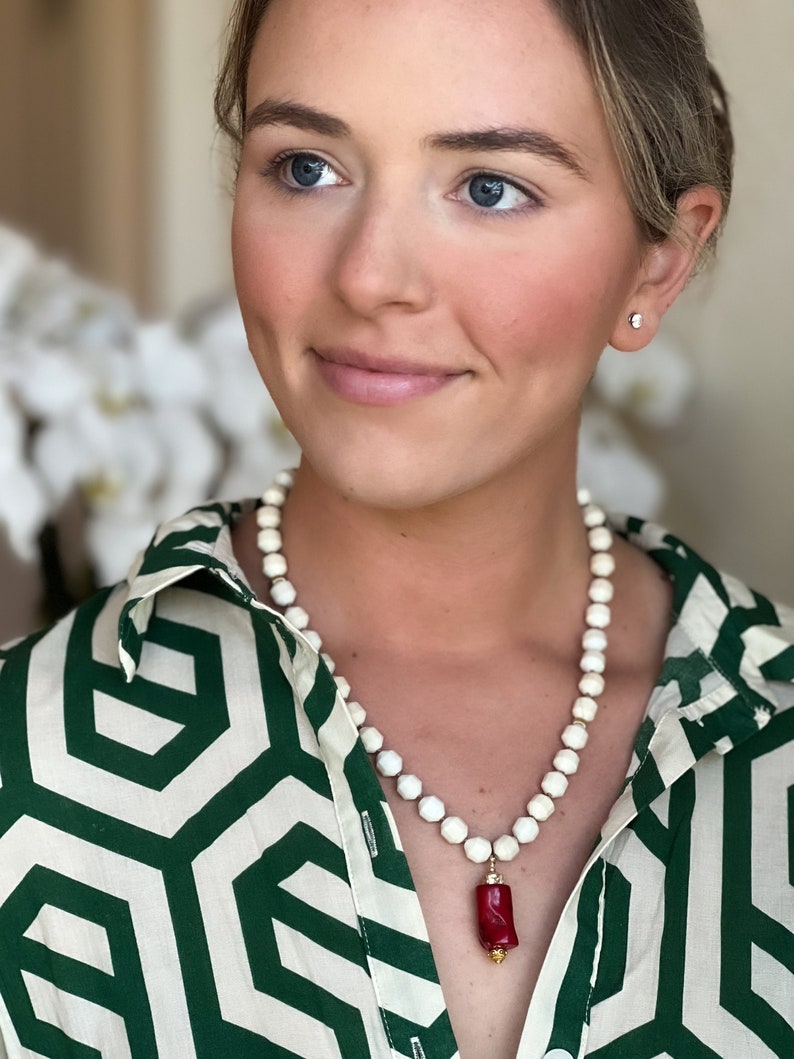 May include: A white beaded necklace with a red coral pendant. The necklace is worn by a person with a green and white patterned shirt.