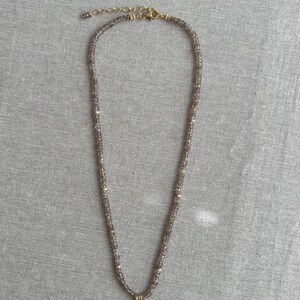 K&ouml;nnte beinhalten: Eine zarte goldene Halskette mit einem kleinen, runden Anh&auml;nger. Die Kette besteht aus winzigen, facettierten Perlen in hellbrauner Farbe.