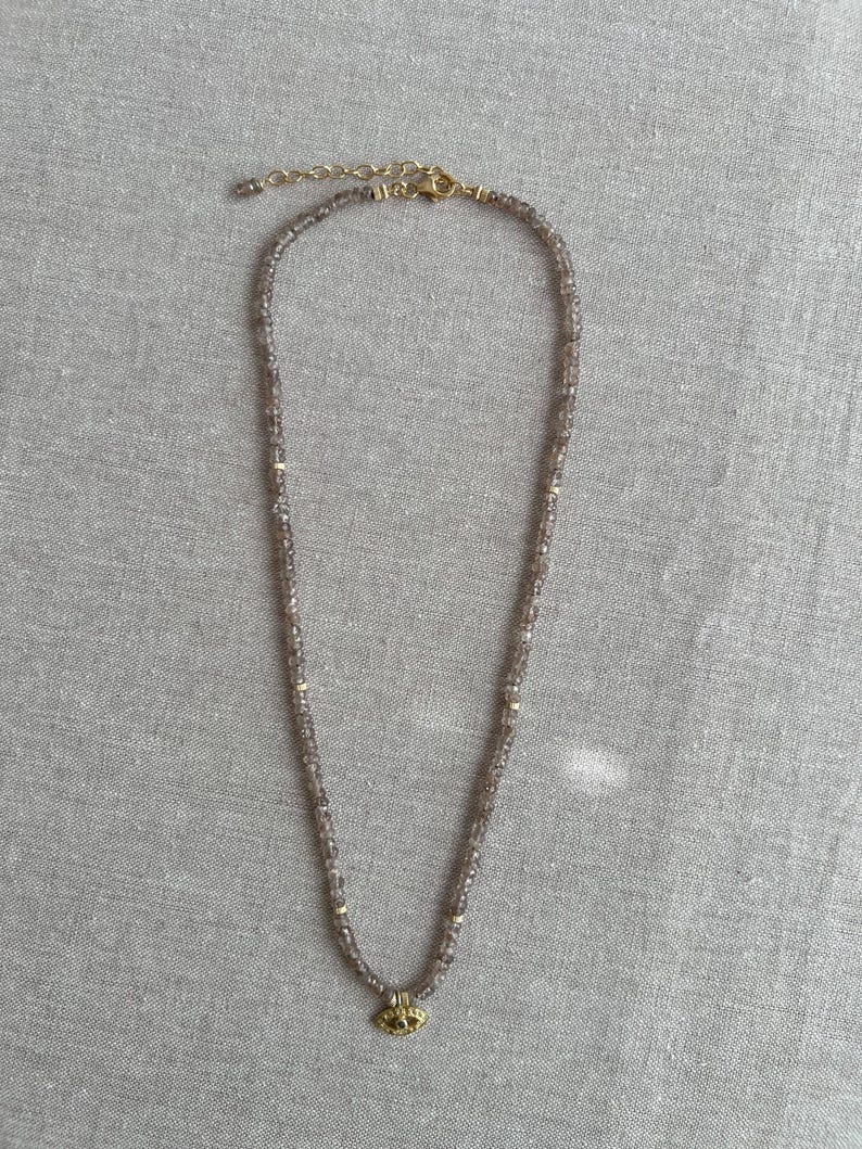 K&ouml;nnte beinhalten: Eine zarte goldene Halskette mit einem kleinen, runden Anh&auml;nger. Die Kette besteht aus winzigen, facettierten Perlen in hellbrauner Farbe.