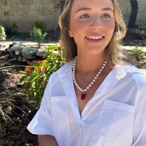 May include: A woman wearing a white button-down shirt with a white and red beaded necklace. The necklace has a red rectangular pendant.