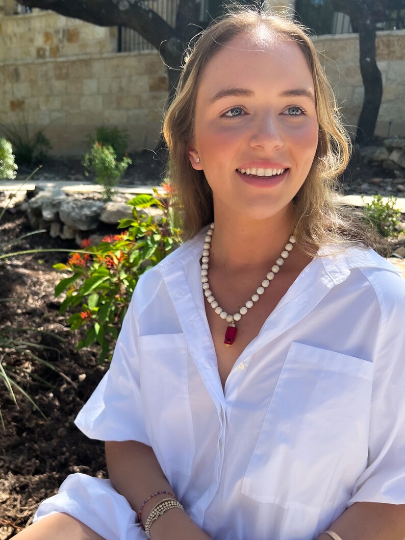 May include: A woman wearing a white button-down shirt with a white and red beaded necklace. The necklace has a red rectangular pendant.