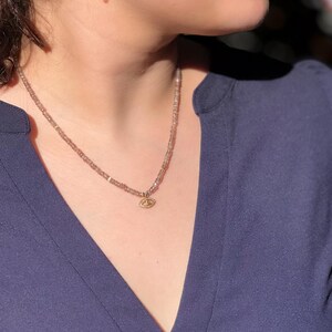 K&ouml;nnte beinhalten: Eine zarte Halskette mit einem kleinen goldenen Augen-Anh&auml;nger. Die Halskette besteht aus winzigen, hellbraunen Perlen und wird von einer Person mit einem dunkelblauen Hemd getragen.