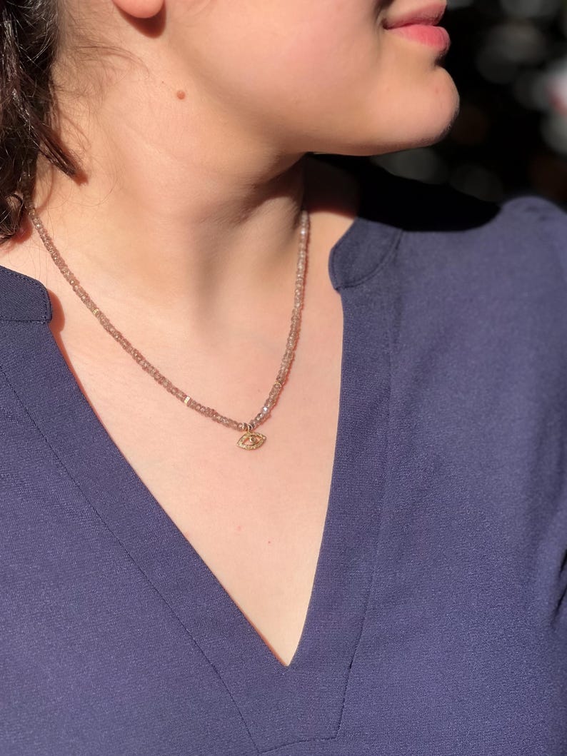 K&ouml;nnte beinhalten: Eine zarte Halskette mit einem kleinen goldenen Augen-Anh&auml;nger. Die Halskette besteht aus winzigen, hellbraunen Perlen und wird von einer Person mit einem dunkelblauen Hemd getragen.