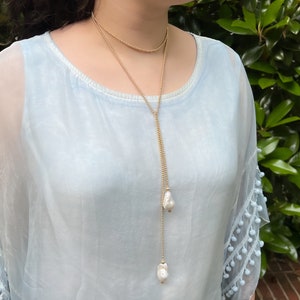 May include: A long gold chain necklace with two large white pearl drops. The necklace is layered over a light blue sheer top with white pom-pom trim.