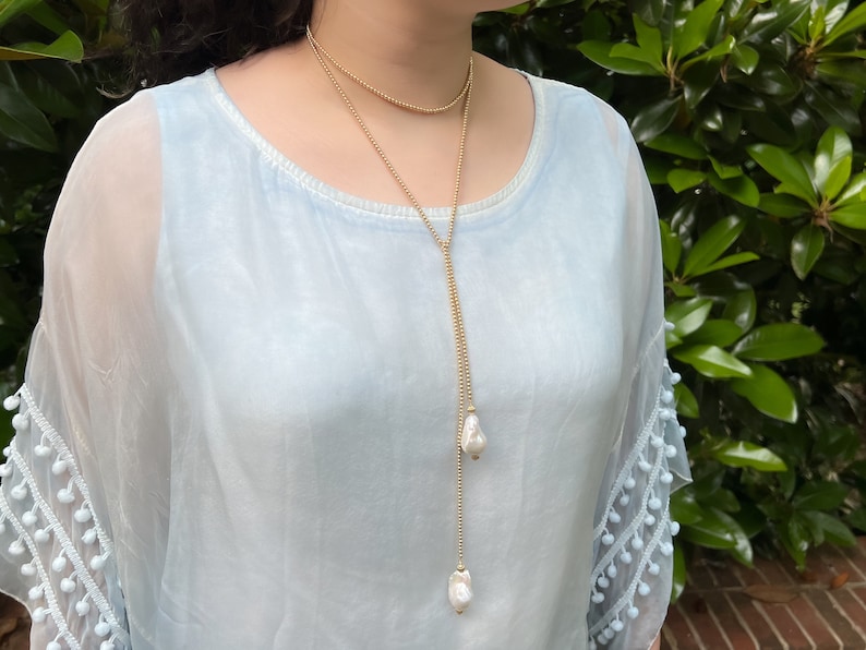 May include: A long gold chain necklace with two large white pearl drops. The necklace is layered over a light blue sheer top with white pom-pom trim.