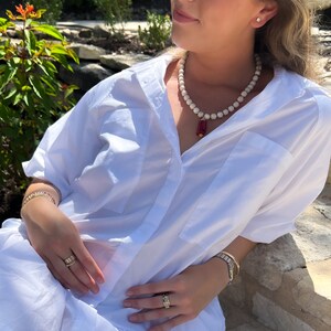 May include: A white button-down shirt with a red and gold pendant necklace. The necklace is made of white beads and has a red rectangular pendant with gold accents.