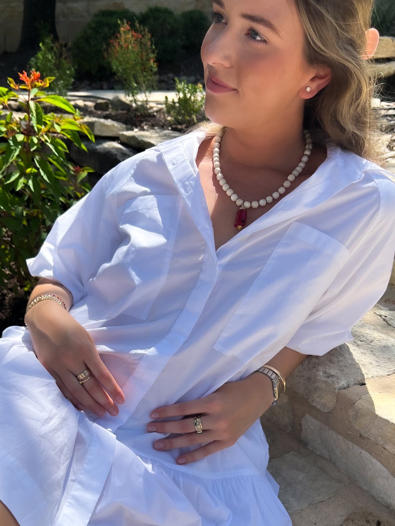 May include: A white button-down shirt with a red and gold pendant necklace. The necklace is made of white beads and has a red rectangular pendant with gold accents.