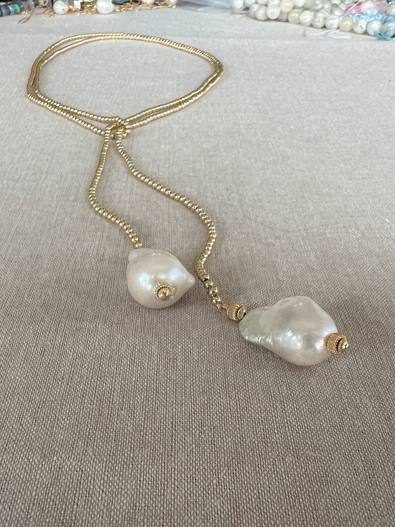 May include: A gold chain necklace with two large white baroque pearls at the ends. The chain is made of small gold beads.