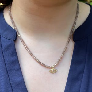 K&ouml;nnte beinhalten: Eine zarte Halskette aus braunen Edelsteinen mit einem goldenen b&ouml;sen Augen-Anh&auml;nger. Der Anh&auml;nger hat einen kleinen roten Edelstein in der Mitte des Auges.