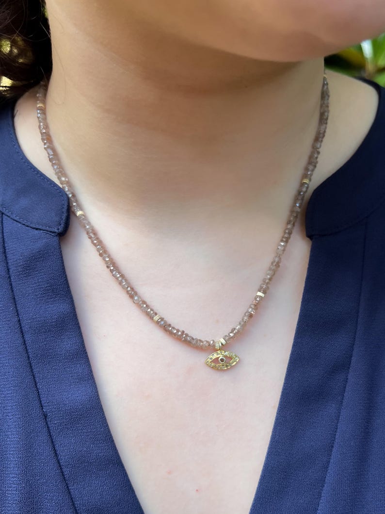 K&ouml;nnte beinhalten: Eine zarte Halskette aus braunen Edelsteinen mit einem goldenen b&ouml;sen Augen-Anh&auml;nger. Der Anh&auml;nger hat einen kleinen roten Edelstein in der Mitte des Auges.