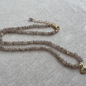 K&ouml;nnte beinhalten: Eine zarte goldene Kettenkette mit einem mehrschichtigen Design aus kleinen, hellbraunen Perlen. Die Kette hat einen goldenen Anh&auml;nger in Form eines Auges mit einem kleinen, klaren Stein in der Mitte.