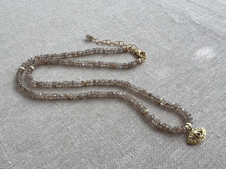 K&ouml;nnte beinhalten: Eine zarte goldene Kettenkette mit einem mehrschichtigen Design aus kleinen, hellbraunen Perlen. Die Kette hat einen goldenen Anh&auml;nger in Form eines Auges mit einem kleinen, klaren Stein in der Mitte.