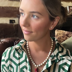 May include: A white and red beaded necklace with a red pendant. The necklace is worn by a person with long brown hair and blue eyes. The person is wearing a green and white patterned shirt.